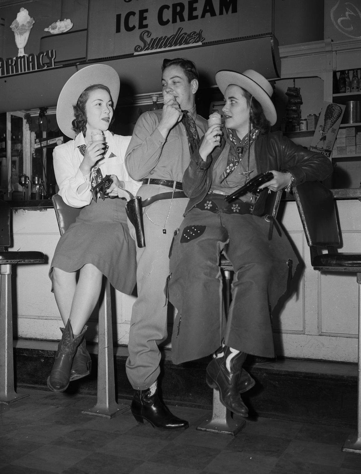Miss Mays and Miss Pray are both wearing cowboy hats, bandannas and cowboy boots and are each holding a handgun. Miss Mays is wearing a blouse and a skirt. Miss Pray is wearing a blouse, coat and trousers. Mr. McFalls is wearing a long-sleeve shirt, a bandanna around his neck, trousers and cowboy boots.