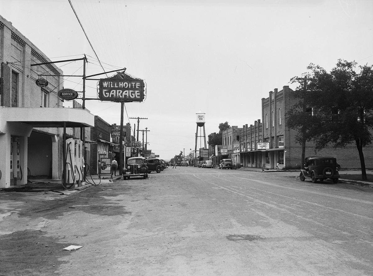 June 1940: View of Grapevine, Texas’, Main Street (Highway 121). On the left side of the road are various businesses, including a gas station, Willhoite Garage, a drugstore and a pharmacy. There are advertisements for Coca-Cola on the buildings. In the background, center, is the Grapevine, Texas, water tower. On the right side of the road is a laundromat and other businesses. The sign outside the laundromat reads, “Help U Self Laundry.”
