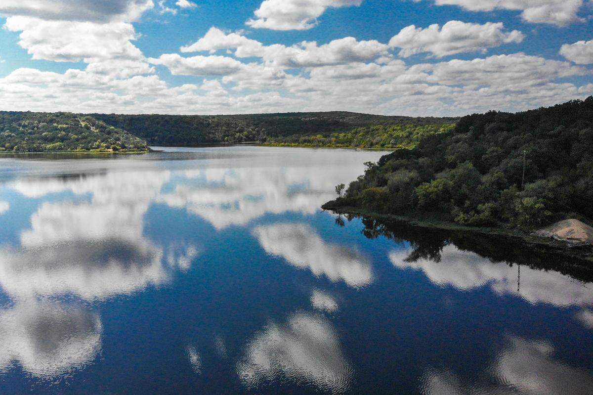 Blue sky and clouds reflected on calm lake waters surrounded by hills full of trees.