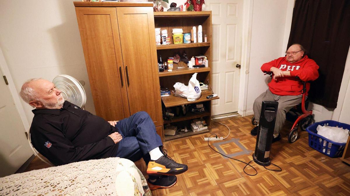 Two men sit across from each other in a small apartment living area. A bed and cabinet can be seen.