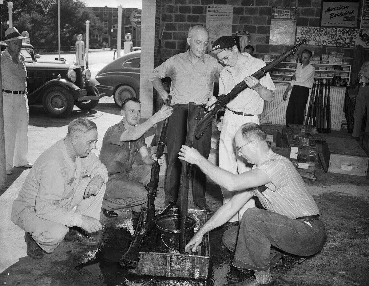 Grogan is the commanding officer of Company B. He is wearing a long-sleeve shirt and trousers. Private Hines, Company A, is dressed in a short-sleeve shirt and trousers. Captain Calder, Company A commander, is wearing a button-up shirt and trousers. Corporal Johnson, Company A, is dressed in a hat, short-sleeve shirt, and trousers. Private Coggins, Company A, is wearing a striped short-sleeve shirt and trousers. There are several unidentified men standing in the background and two automobiles are parked outside the garage.