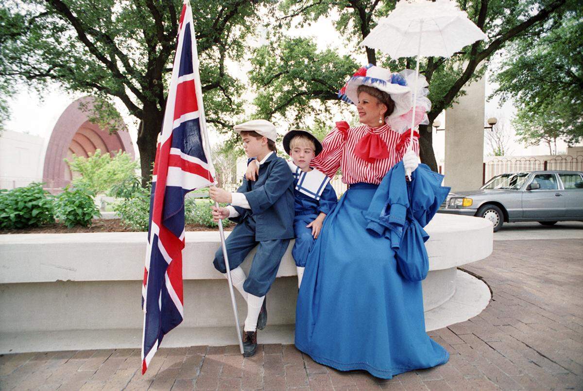 A woman and two boys wearing red, white and blue costumes similar to the 1800s sit on an outside bench while one boy holds a large British flag. 