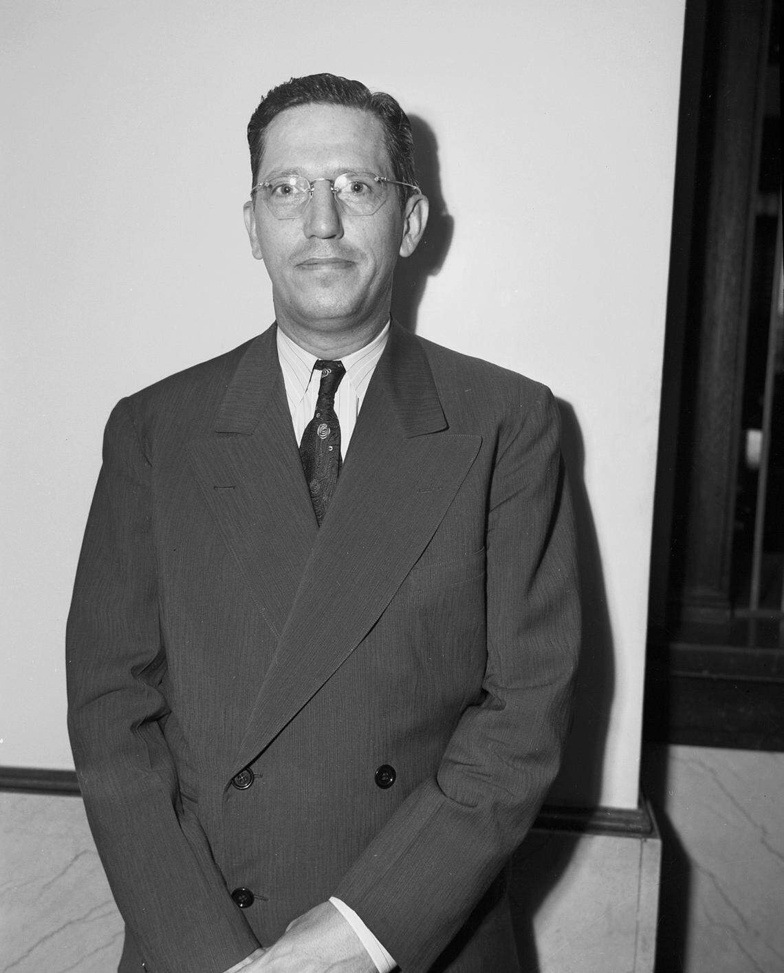 A black and white photo of a man wearing a suite in front of a white wall.