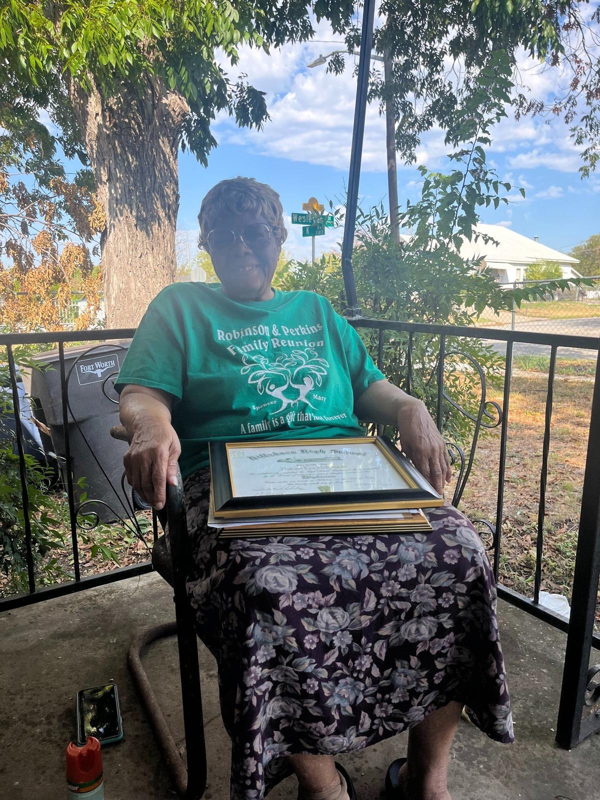 A woman wearing a green T-shirt and a floral skirt sits in a chair on a front porch. On her lap, there is a stack of framed certificates and diplomas. 