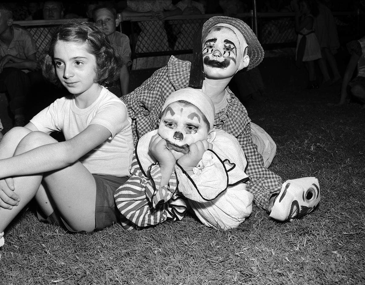 Three kids sit on the ground, two of them dressed as clowns, appearing with serious expressions.
