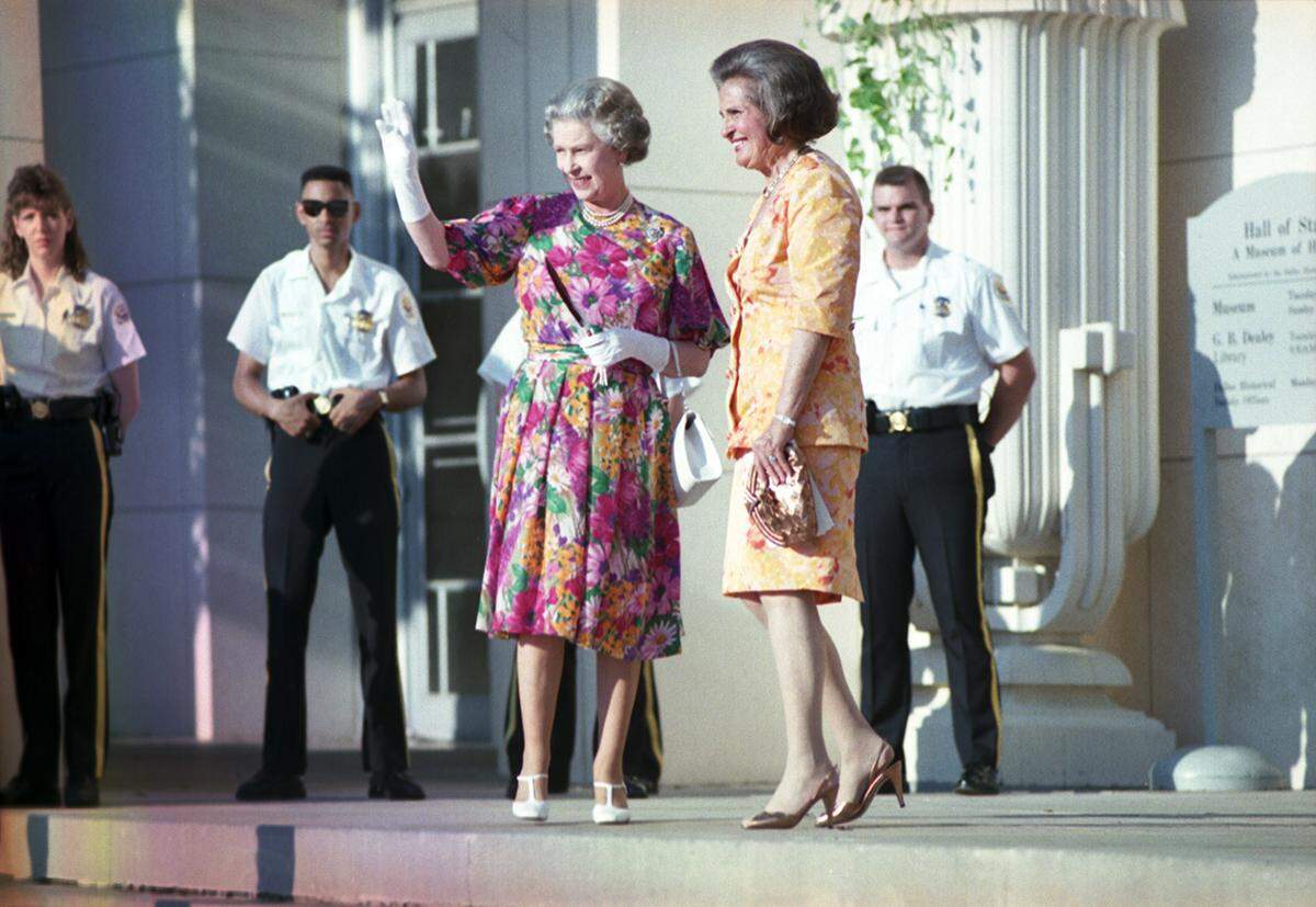 Queen Elizabeth in a pink floral dress and white gloves waves to a crowd as she stands next to the smiling mayor of Dallas, Annette Straus, with security standing at attention behind them. 