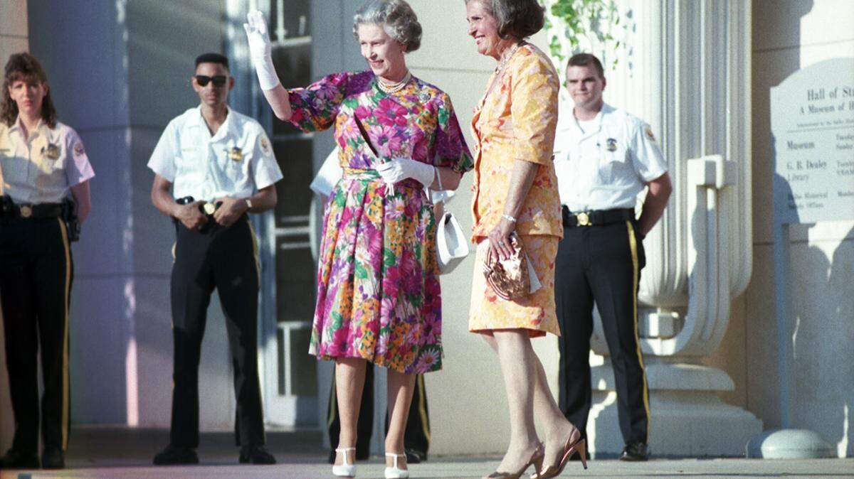 Queen Elizabeth in a pink floral dress and white gloves waves to a crowd as she stands next to the smiling mayor of Dallas, Annette Straus, with security standing at attention behind them. 