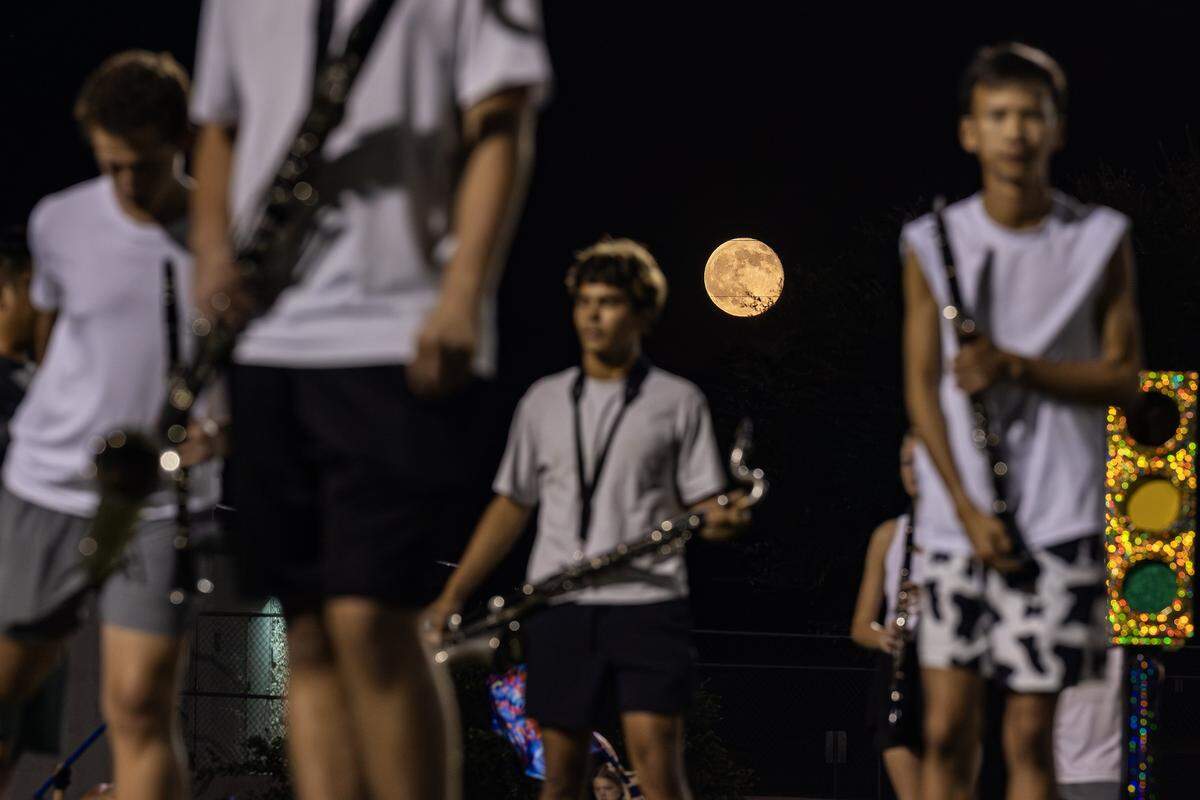 A large orange full moon can be seen in a dark sky while in the foreground stand woodwind players in gym attire. 