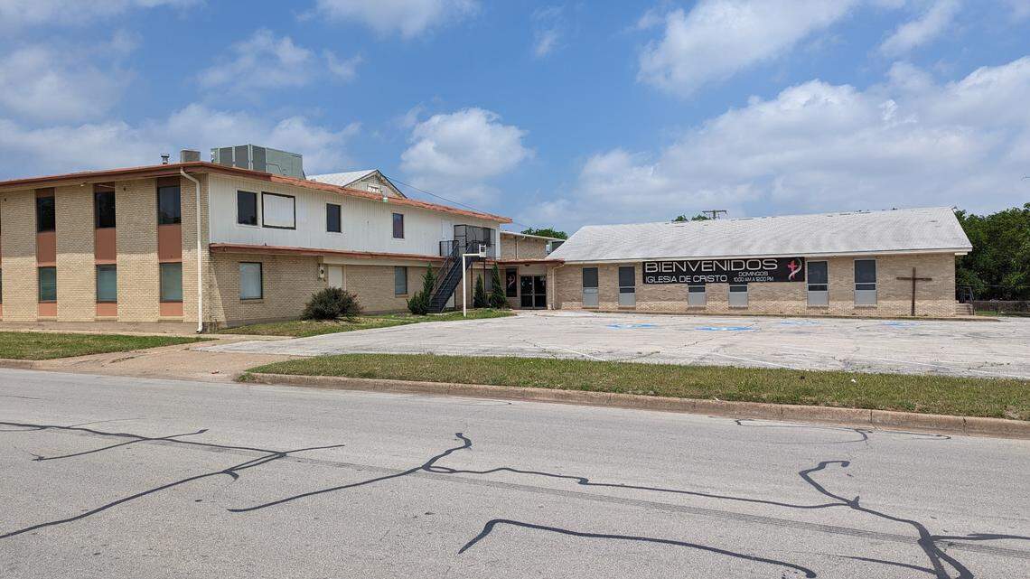 A two story building to the left with a parking lot in the middle and an A-frame house behind it. 