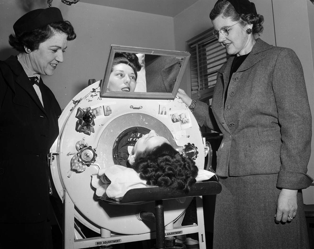 Evelyn Marie DeBevoise, flanked by Gussie Lee Hellman (left) and Elizabeth Rumph (right), talks about photos of her family posted on her iron lung door. DeBevoise, hospitalized at City-County Hospital (now JPS), contracted polio in 1952 at age 25 before vaccines were developed.