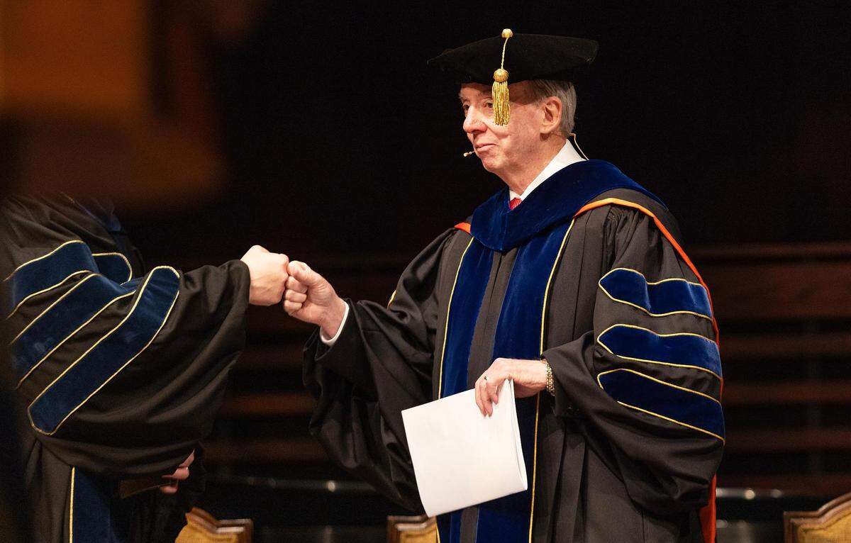 A man in professor cap and gown on the right fist bumps another person who is obscured on the left.