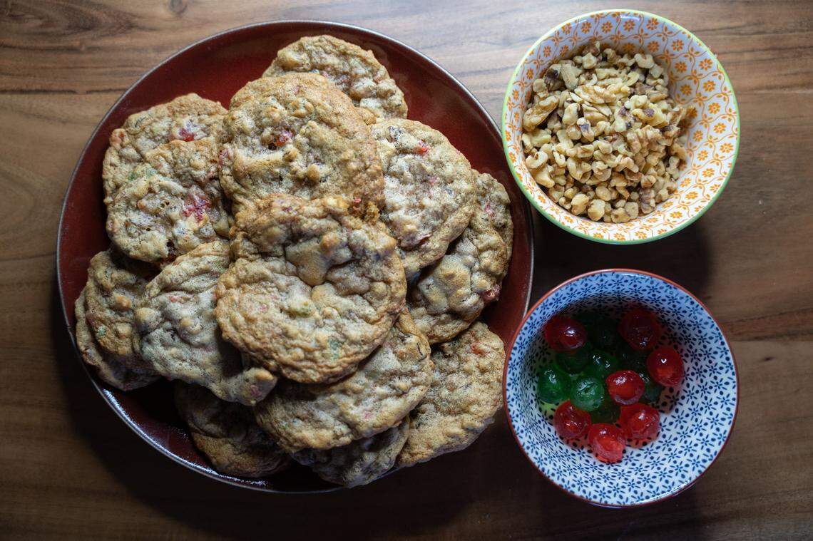 Fruitcake cookies pictured in a stack alongside green and red cherries and crushed nuts.