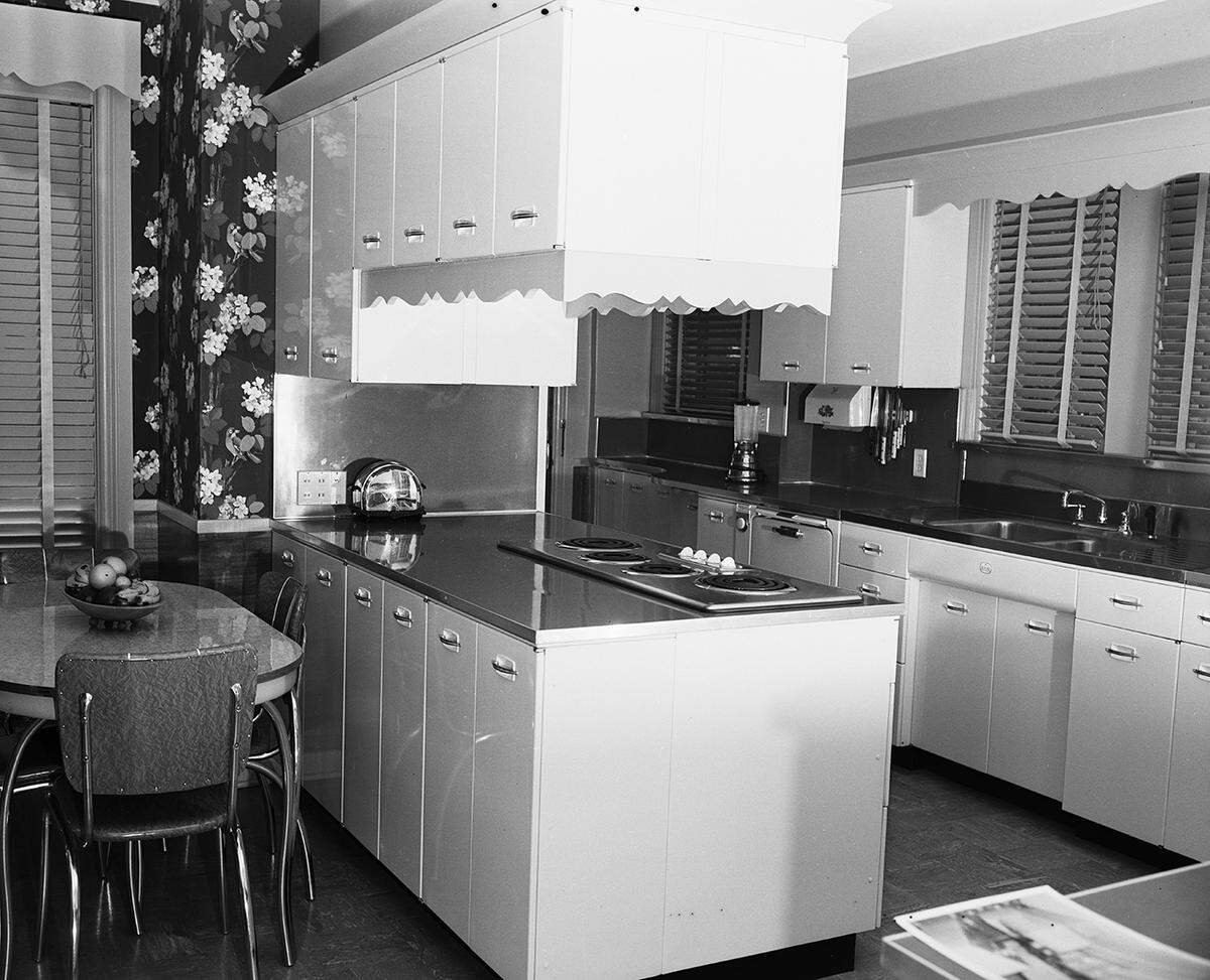 April 23, 1951: The home of Dr. and Mrs. Tom Black, 2600 Ryan Place Drive. At the left is the breakfast area, furnished with metal chairs and table with red plastic top. The peninsula jutting out in the center of the room shows cabinet space built around the range. The color scheme is sage green, white and red. Dogwood blossoms appear in the green wallpaper. The floor is gray rubber tile. Chair-height wainscoting of Philippine mahogany was used around the open walls.