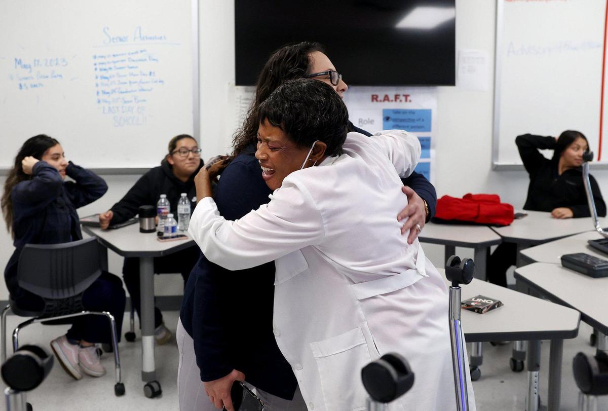 A woman in a white lab coat smiles while hugging Ramsey in a classroom with students seated behind them.