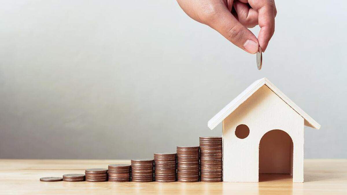 A hand drops a penny in a the roof of a toy house, like a piggy bank. Pennies are stacked in seven increasingly taller piles to the left of the house. 