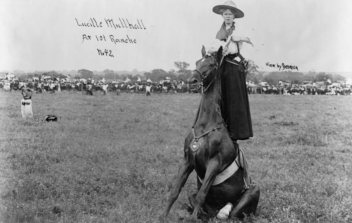 Expert rider Lucille Mulhall is photographed standing on the back of a horse.