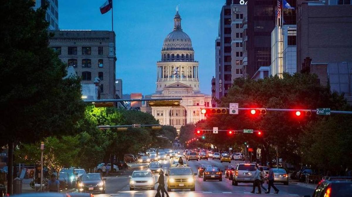 The Texas State Capitol building stands past cars and pedestrians crossing Congress Avenue at dusk in Austin.