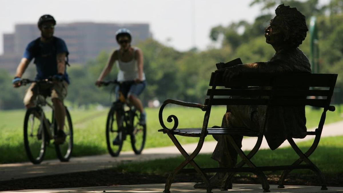 A bronze sculpture of Mark Twain reading his book Huckleberry Finn watches bike riders roll on the trail at Trinity Park.