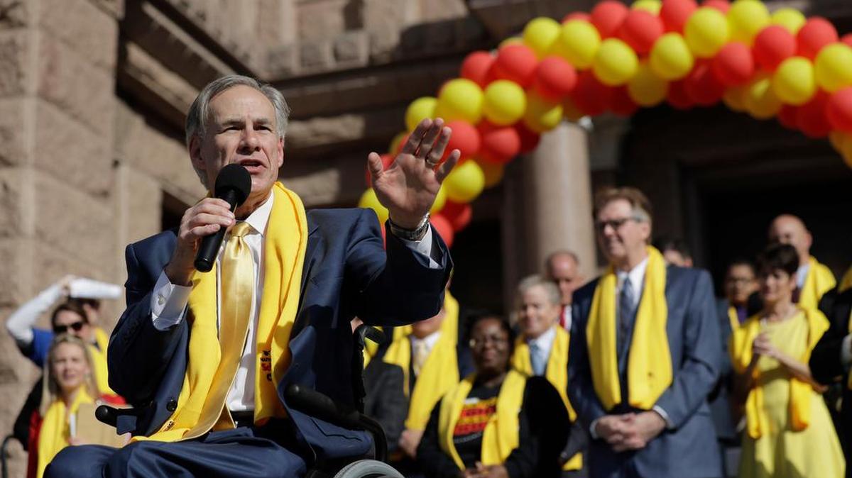 Gov. Greg Abbott gestures while speaking to a crowd. He's wearing a blue suit and a yellow scarf, and is seated in a wheelchair in front of an arc of red and yellow balloons. 