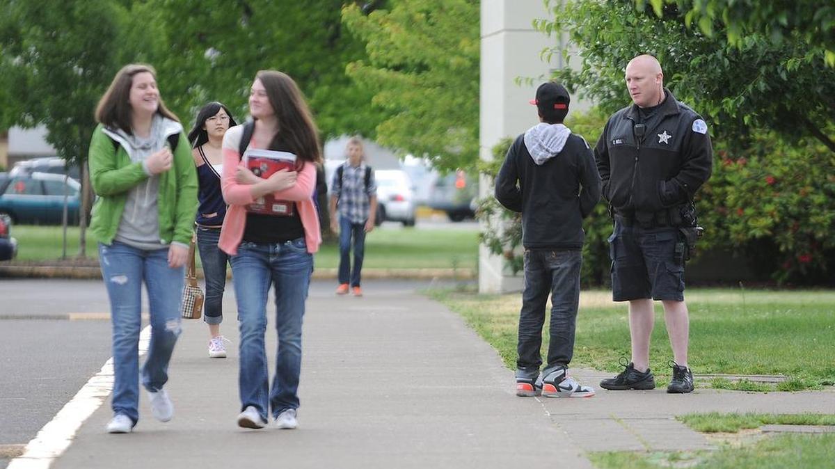 A police school resource officer talks with a high school student while greeting teenagers as they arrive at school.