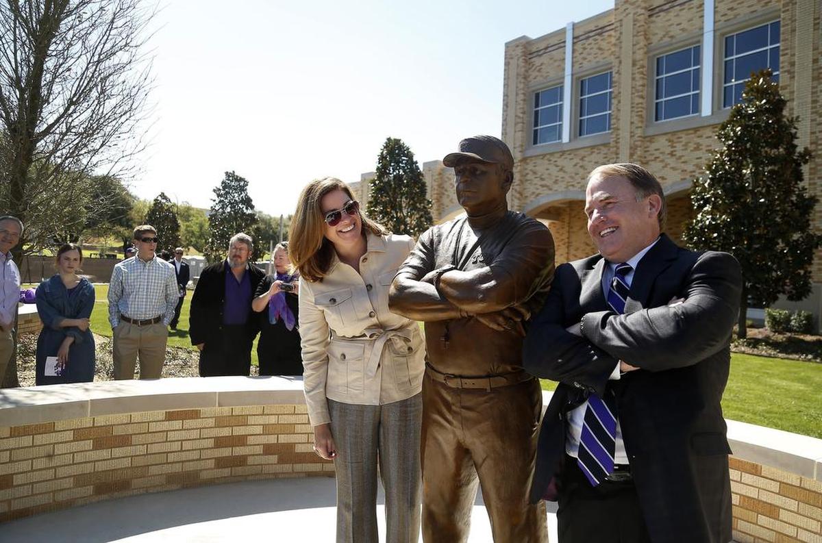 A woman peaks around a bronze statue to smile at Gary Patterson, wearing a suit with arms crossed.